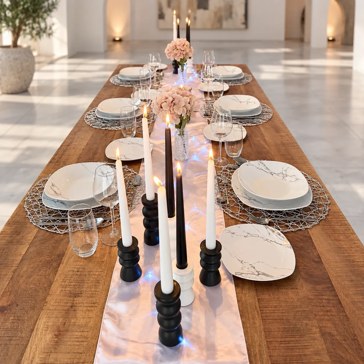 Lange houten tafel met een glanzende satijnen loper met lichtjes, vierkant servies in marmerlook op zilveren opengewerkte placemats, witte en zwarte kaarsen op zwarte en witte keramische kandelaars en perzikkleurige hortensia's in glazen vaasjes