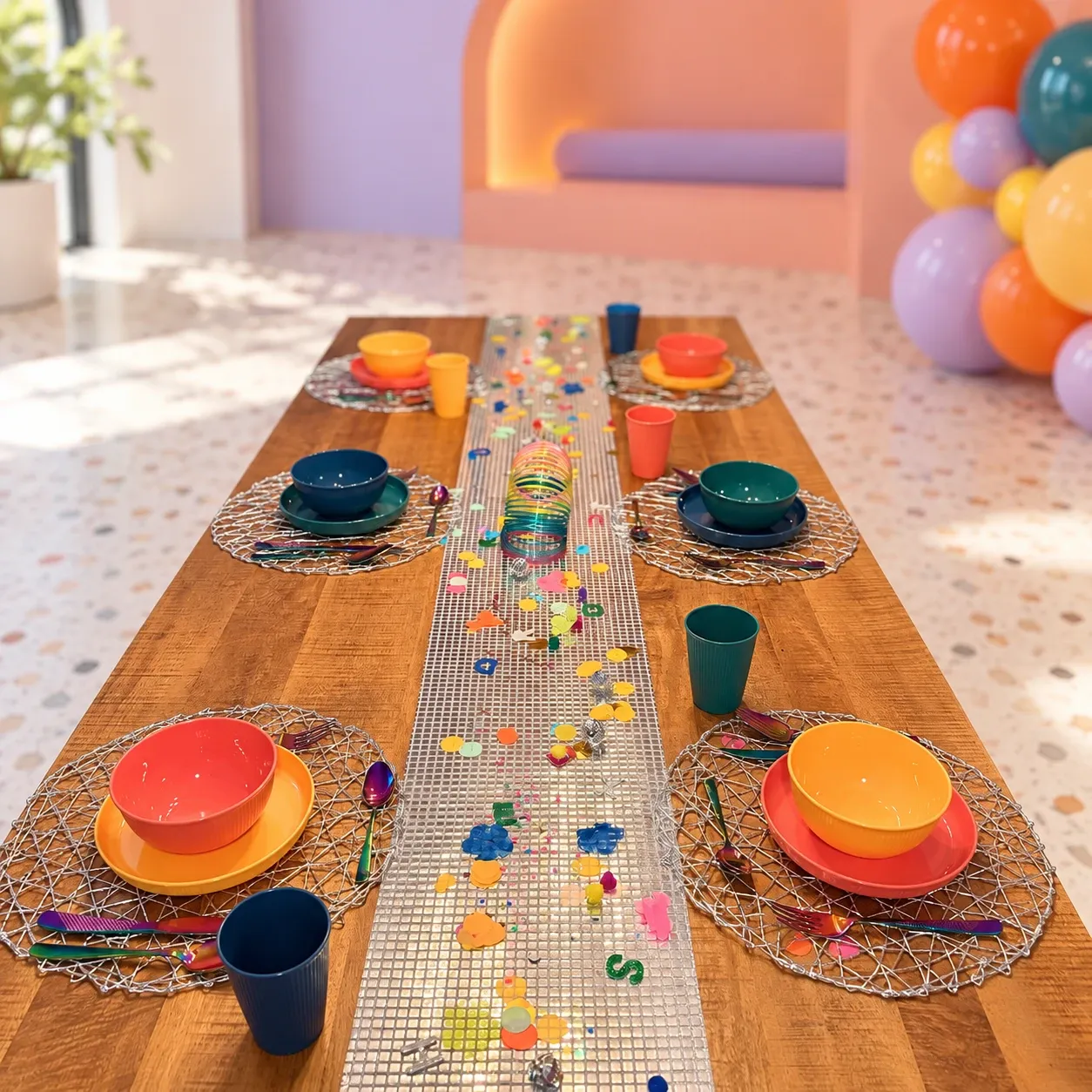Houten tafel met een zilveren mozaïekloper vol confetti, geribbelde kinderborden, kommen en bekers in oranje, geel, groen en blauw op zilveren placemats, en regenboogbestek