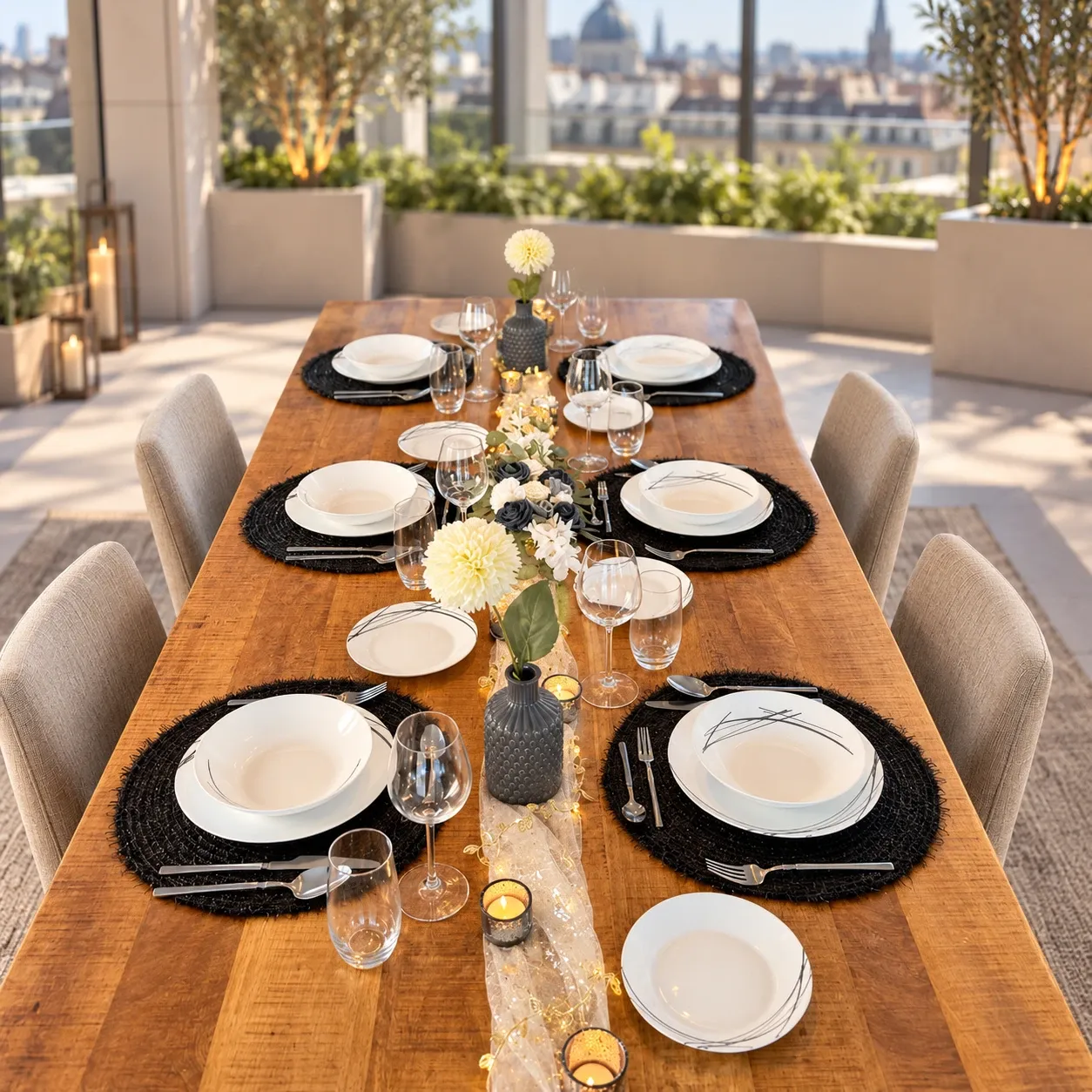 Lange houten tafel op een dakterras met wit servies met zwarte lijnen op zwarte ronde placemats, zilver bestek, een goudkleurige doorschijnende loper met lichtjes, witte bolchrysanten in grijze vaasjes, een bloemstuk van witte en donkere rozen en waxinelichtjes in rookglas